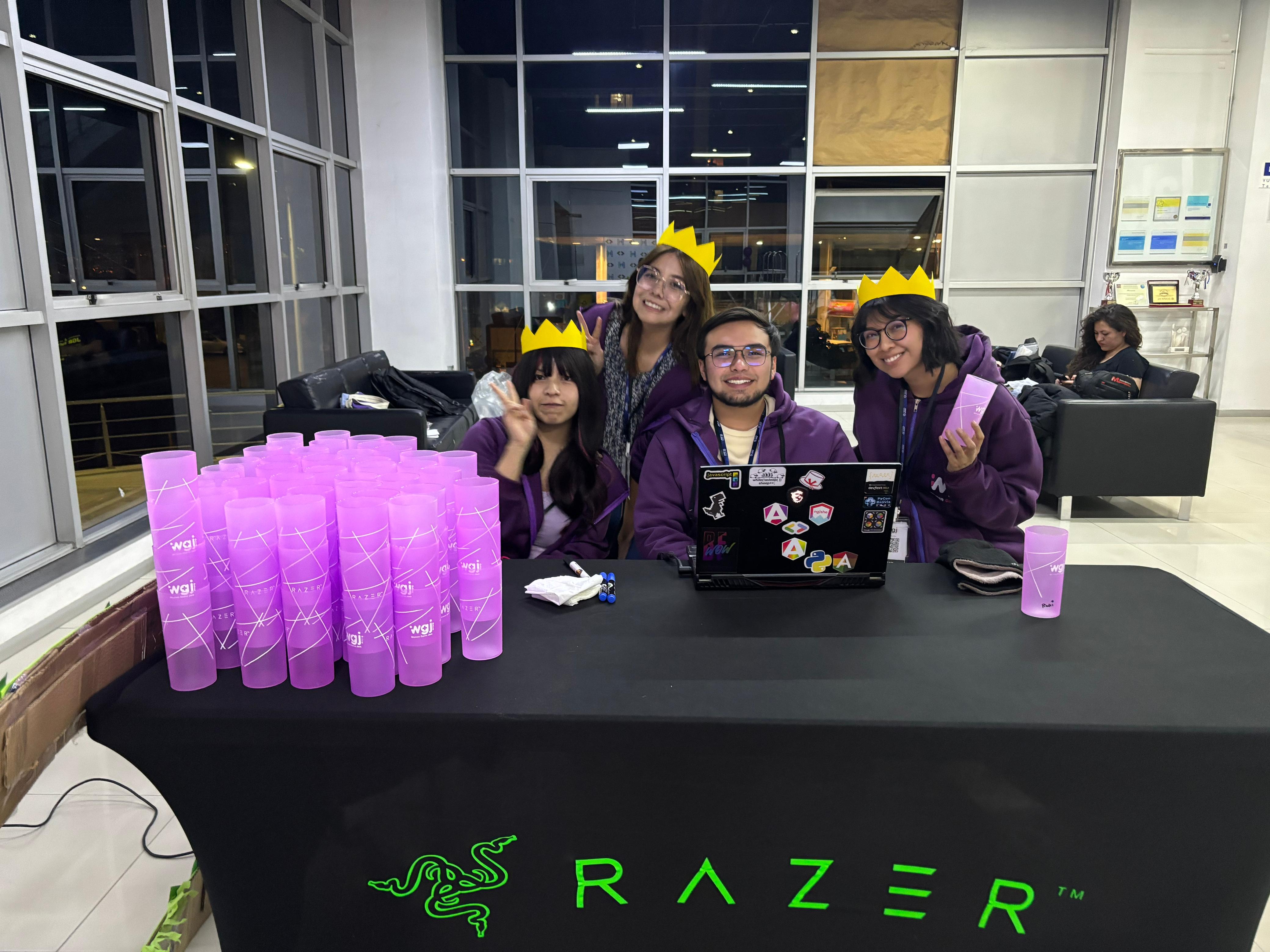 Equipo organizador de la Women Game Jam con coronas de papel junto a vasos morados WGJ en la mesa de registro